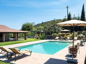 A swimming pool with lounge chairs and umbrellas, surrounded by a landscaped garden and a hill in the background.