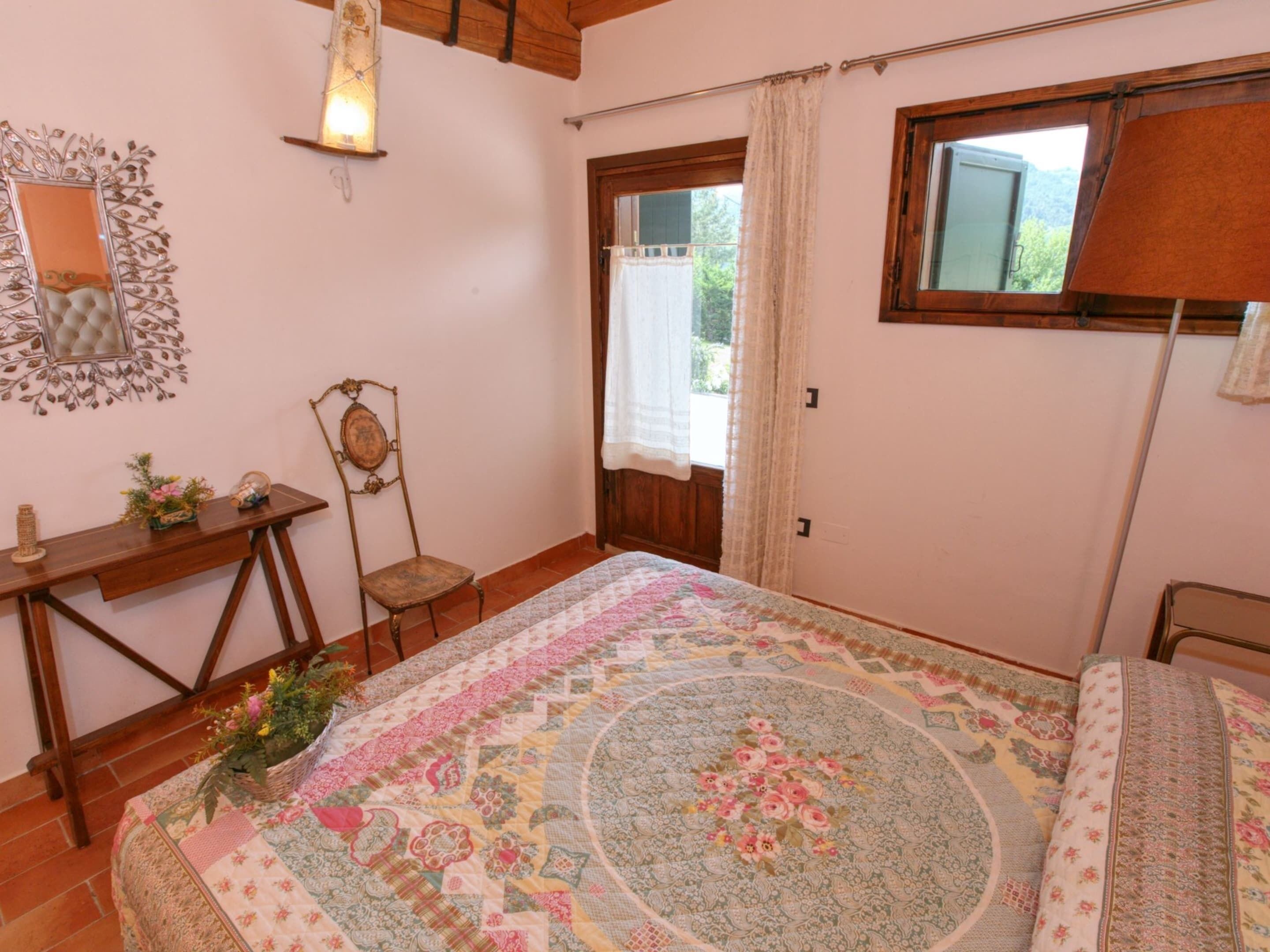 A bedroom with a large bed, a wooden desk, a chair, a mirror, and a window with curtains.
