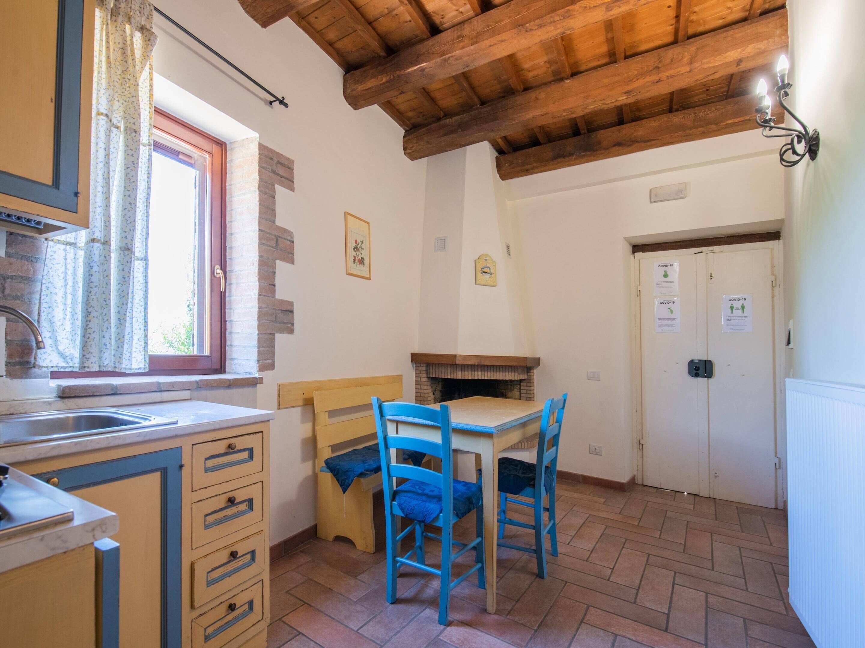 Una pequeña cocina con una mesa y sillas de madera, una ventana con cortinas, y una puerta con señalización de COVID-19.
