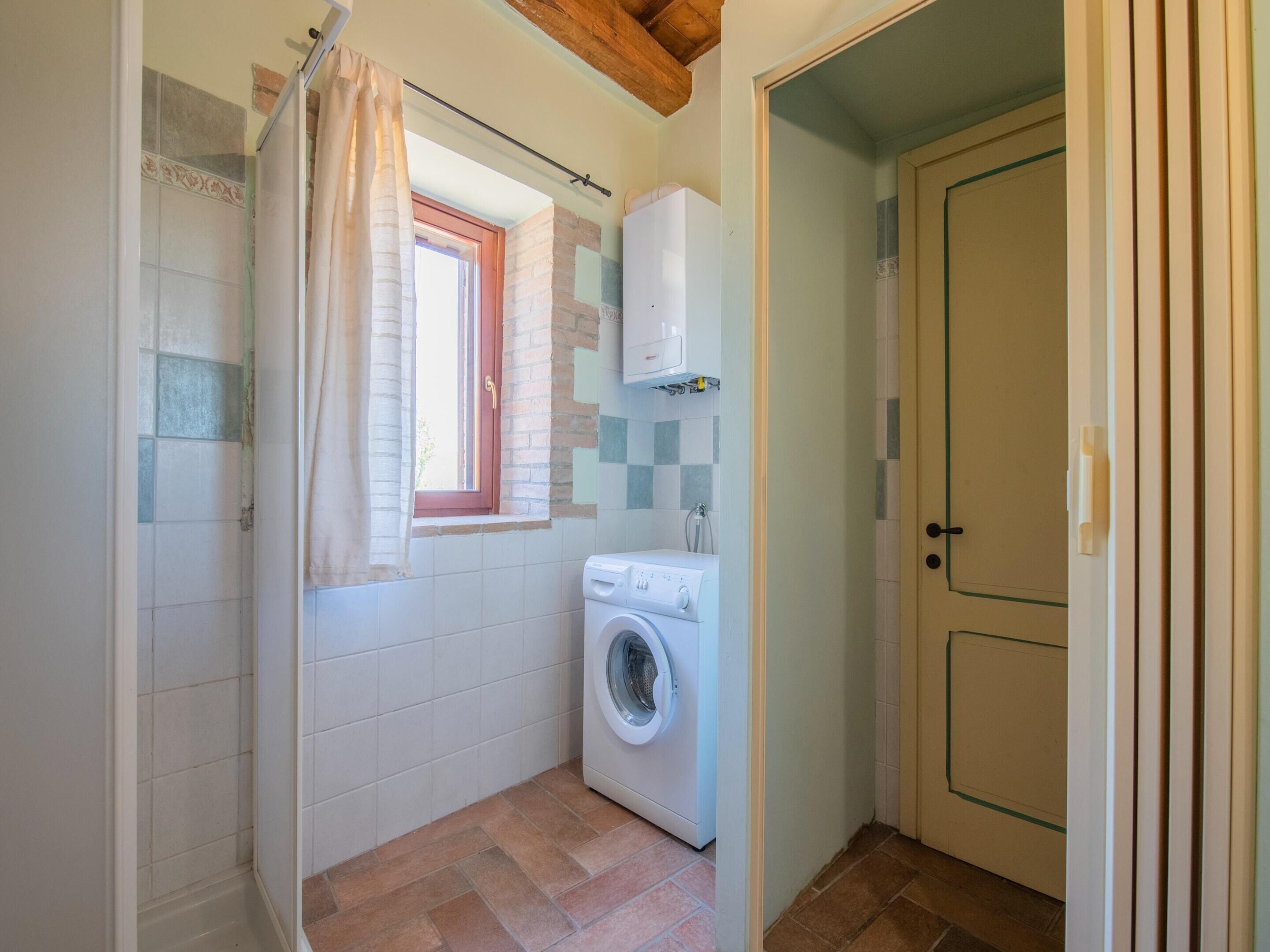A compact bathroom with a washing machine, a shower with a curtain, and a window with a red frame.