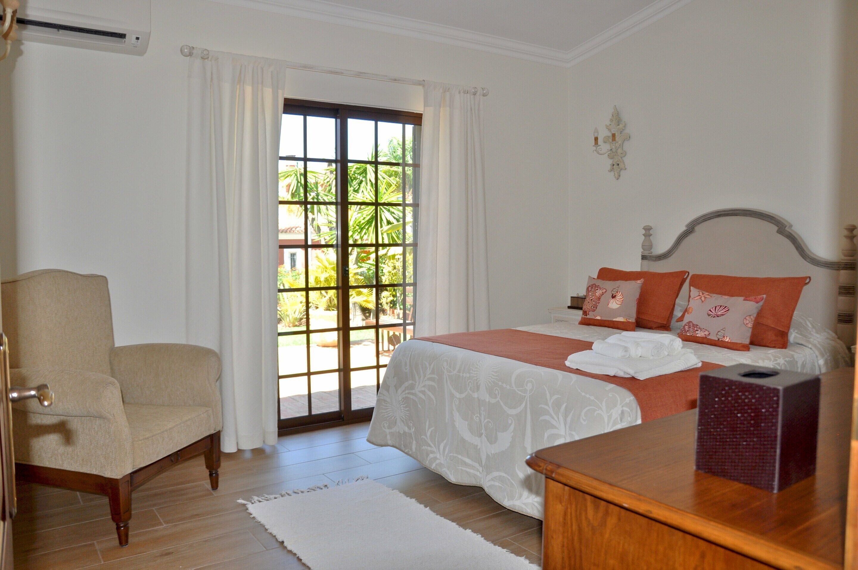 A bedroom with a bed, a chair, a bedside table, and a window with a view of greenery.