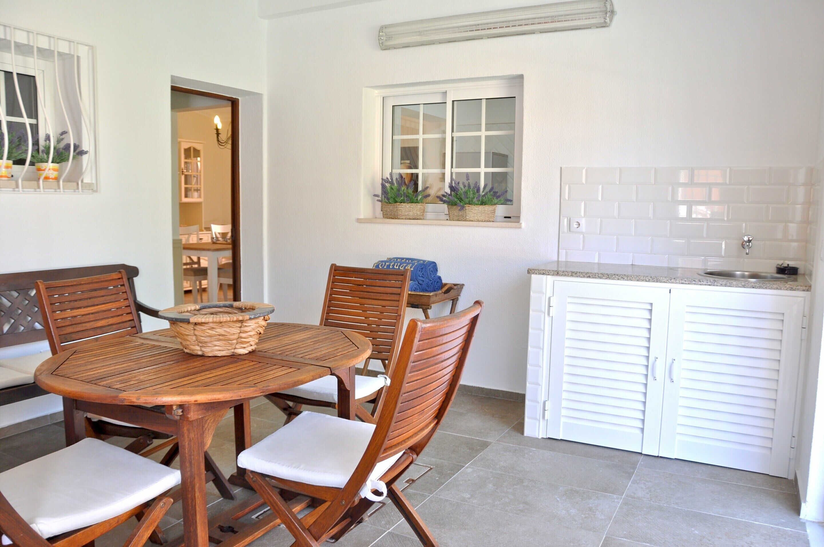 A dining area with a round wooden table and chairs, a small kitchen with white cabinets, and a window with potted plants.