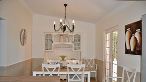 A dining area with a wooden table and white chairs, a chandelier, and a window with a view of greenery. - Villa in Quiet Area with Private Pool near Golf Courses & Marina (Loulé)