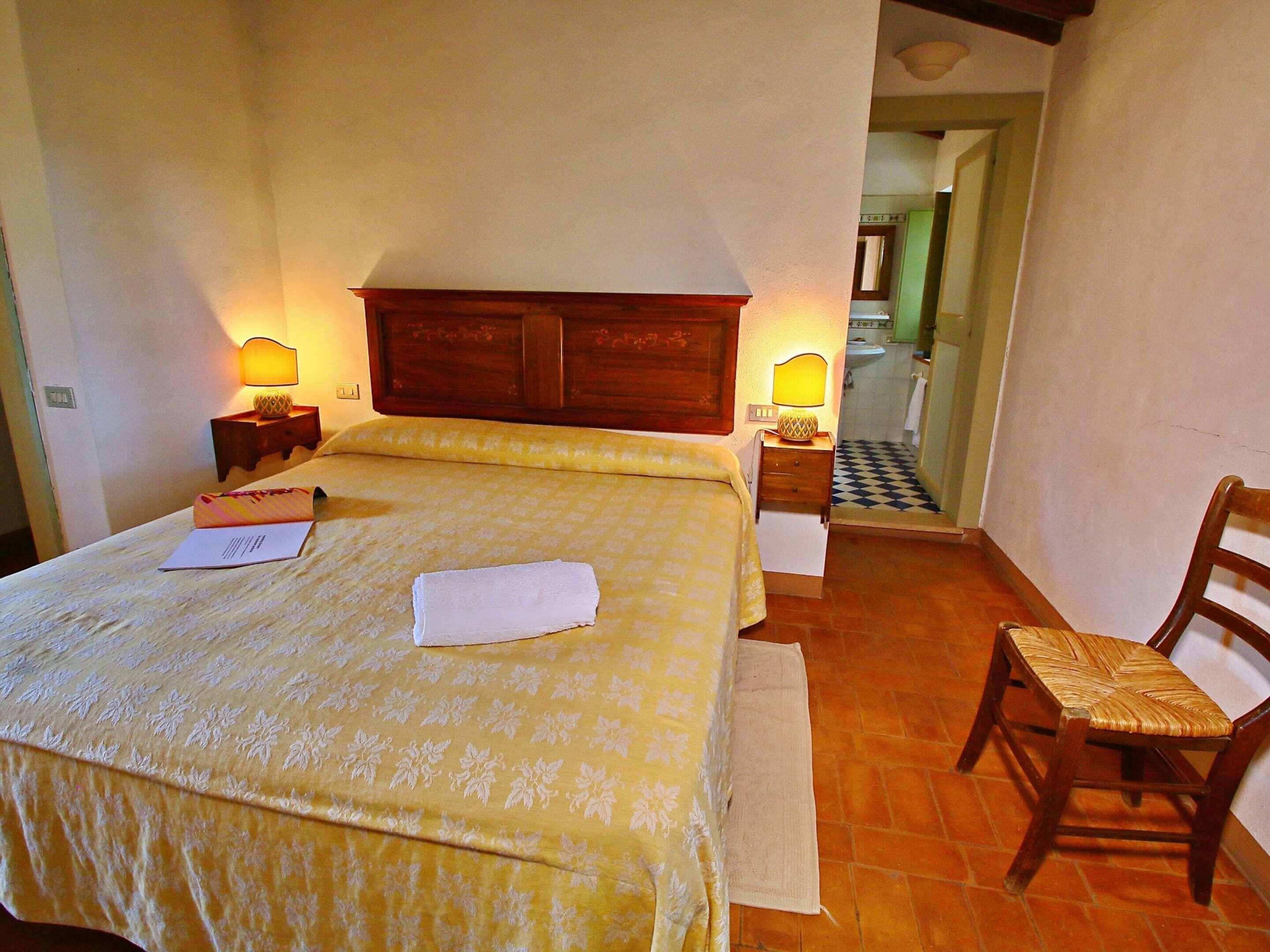 Une chambre d’hôtel avec un lit, deux lampes de chevet, une chaise en bois et une salle de bain visible à travers une porte ouverte.