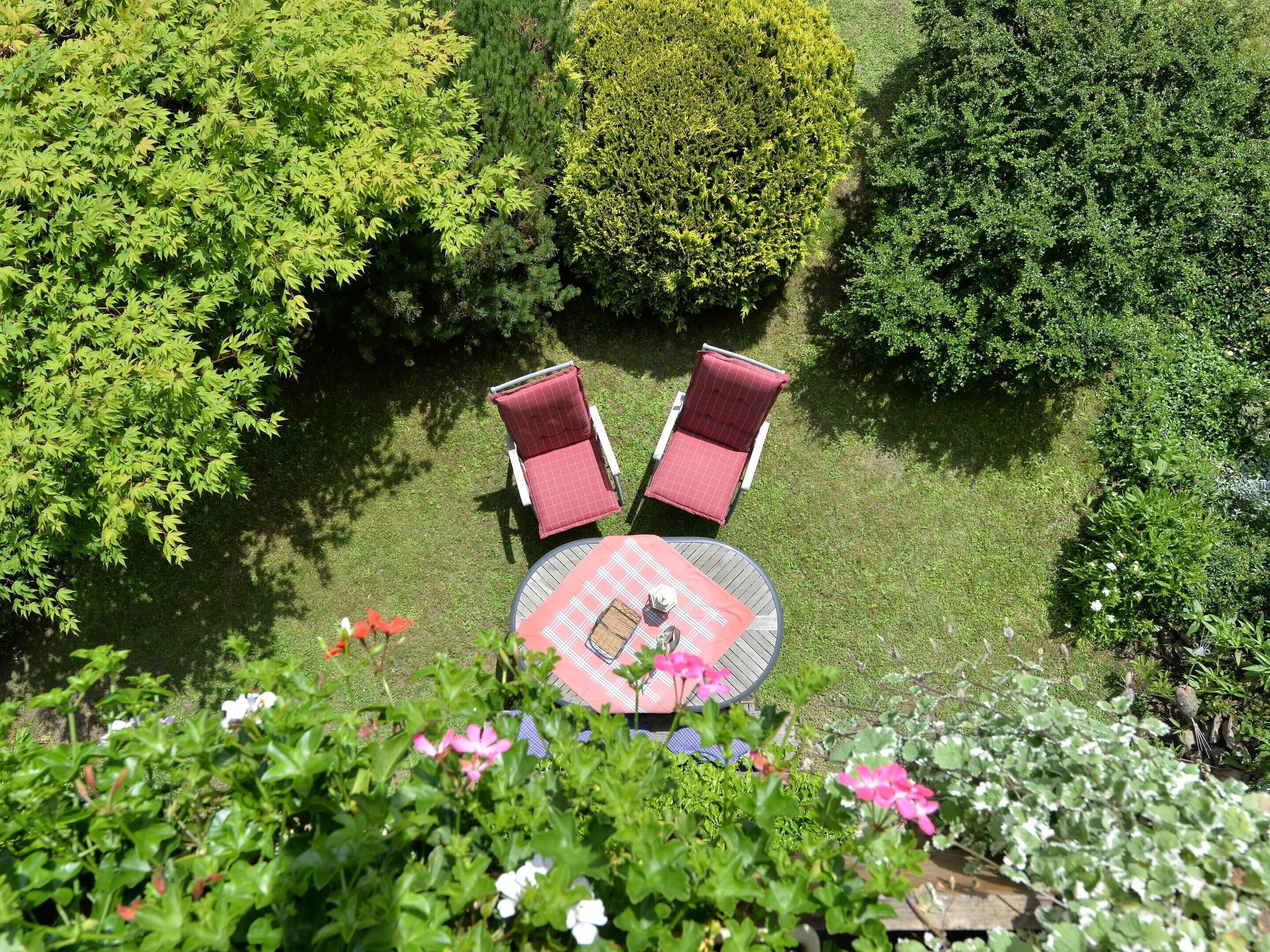 Ein Sitzbereich im Freien mit zwei roten, gepolsterten Stühlen und einem runden Tisch, umgeben von üppigem Grün und blühenden Pflanzen.
