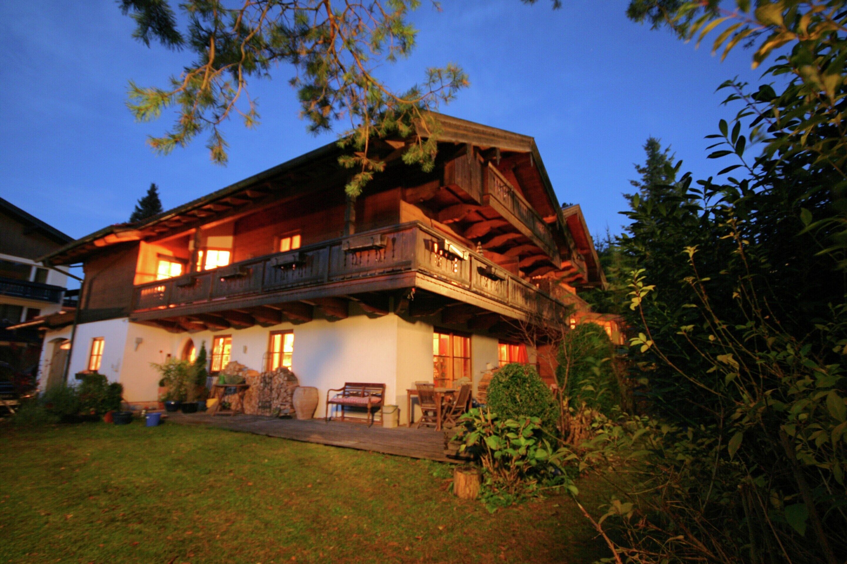 A two-story wooden house with a balcony, surrounded by greenery.