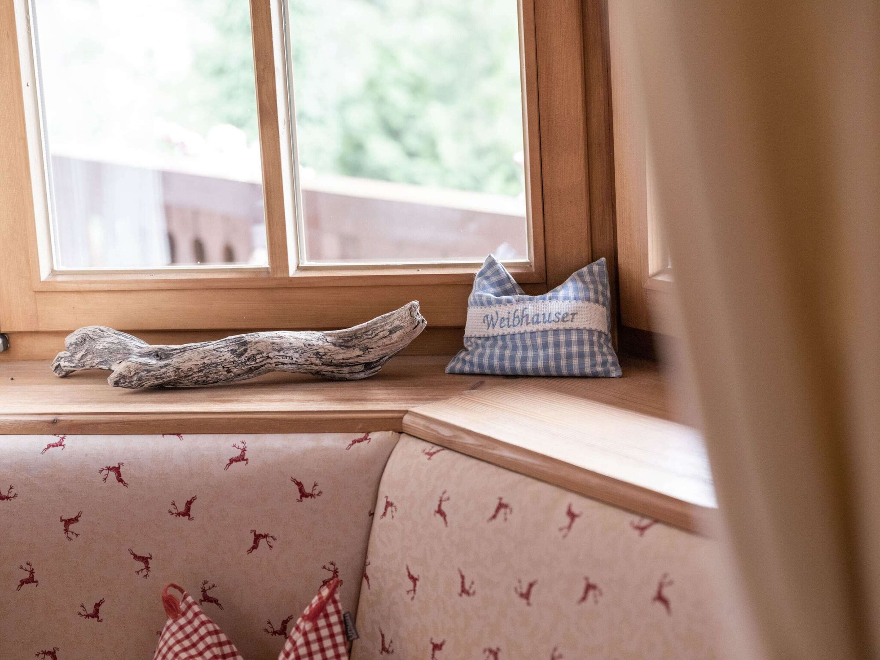 Asiento junto a la ventana con cojín de cuadros, almohada decorativa con la palabra "Weibhausler" y un trozo de madera a la deriva.