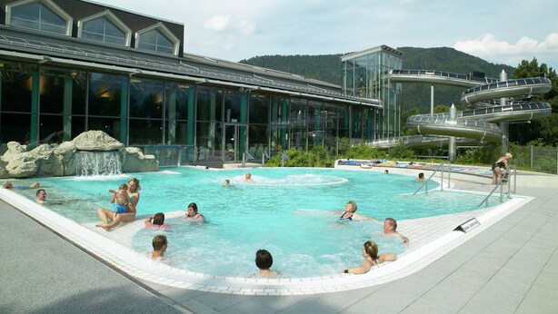 Zona de piscina con un edificio de cristal, tobogán de agua y montañas al fondo.
