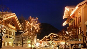 Una calle nevada con edificios decorados con luces, un árbol en el centro y una montaña al fondo.
