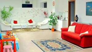 Un salon avec des canapés blancs, un canapé rouge, une table basse, un tapis, une plante et un espace de jeux pour enfants.