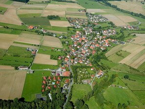 Luchtbeeld van een landelijk dorp, met huizen gegroepeerd langs een hoofdweg, omringd door velden en groen.
