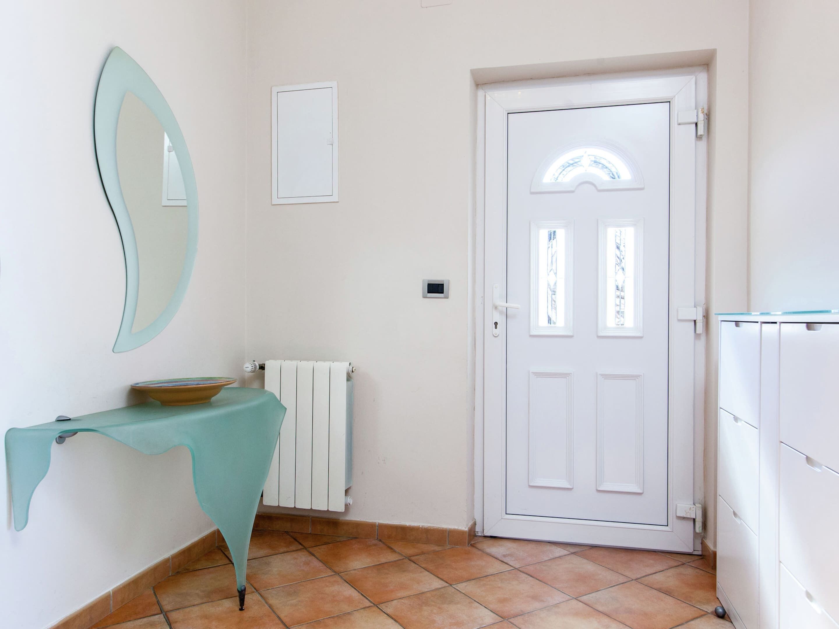 Un passadís amb una porta blanca, un radiador, un mirall fixat a la paret i una taula amb un bol.