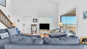 A spacious living room with a large gray sofa, a wooden chair, a TV on a stand, and a window with blinds.