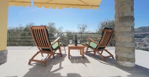Deux fauteuils de jardin en bois avec des coussins verts, une petite table en bois et une bouteille de vin sur un balcon agrémenté d’un pilier en pierre, offrant une vue sur la ville et les collines.