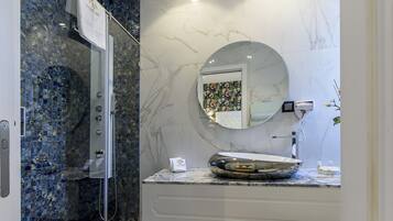 Un baño con cabina de ducha de vidrio, lavabo de mármol y un espejo redondo, además de un mueble de tocador blanco.