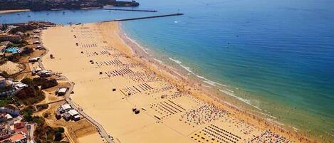Vista aérea de una playa con filas de tumbonas y sombrillas, un muelle que se adentra en el agua y edificios a lo largo de la costa.