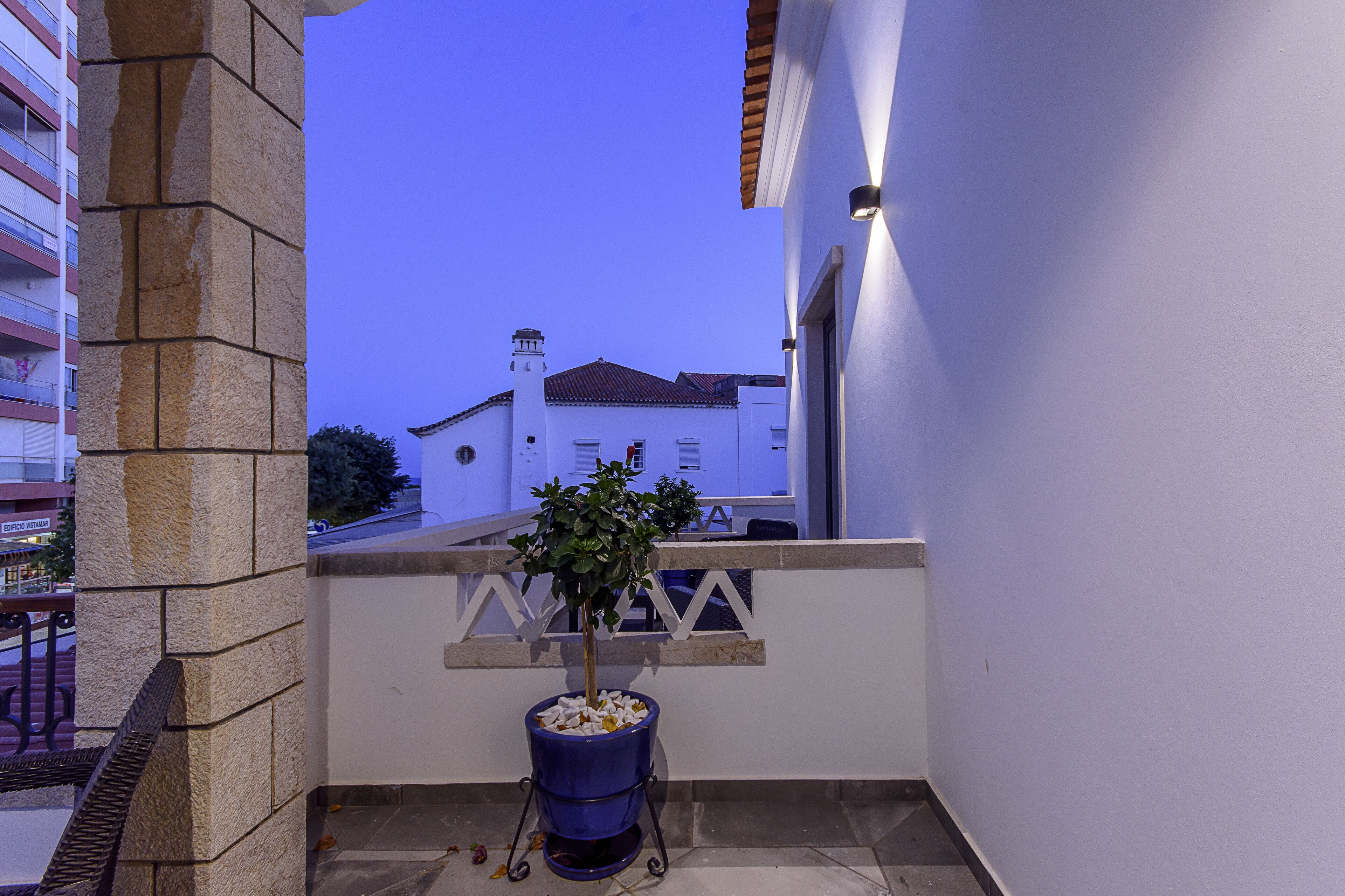 A balcony with a potted plant, a stone pillar, and a view of a building with a tiled roof.