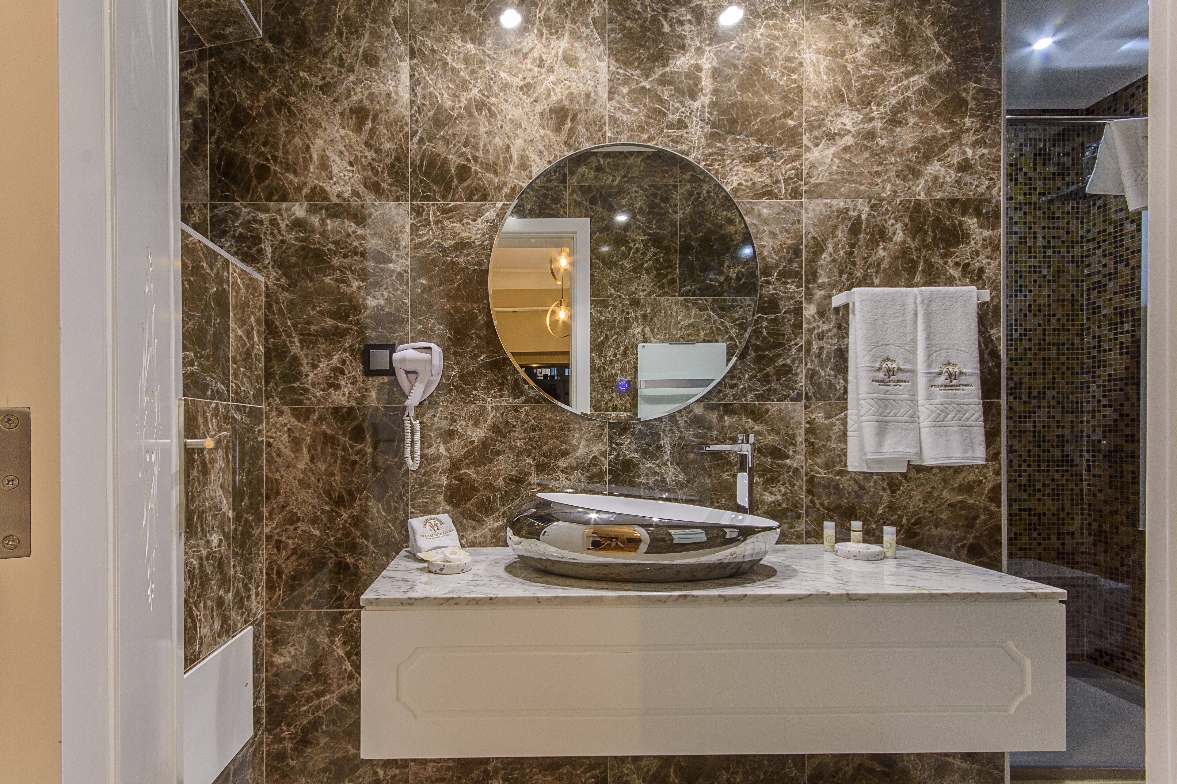 A bathroom with a marble wall, a round mirror, a sink with a vessel basin, and a towel rack with towels.