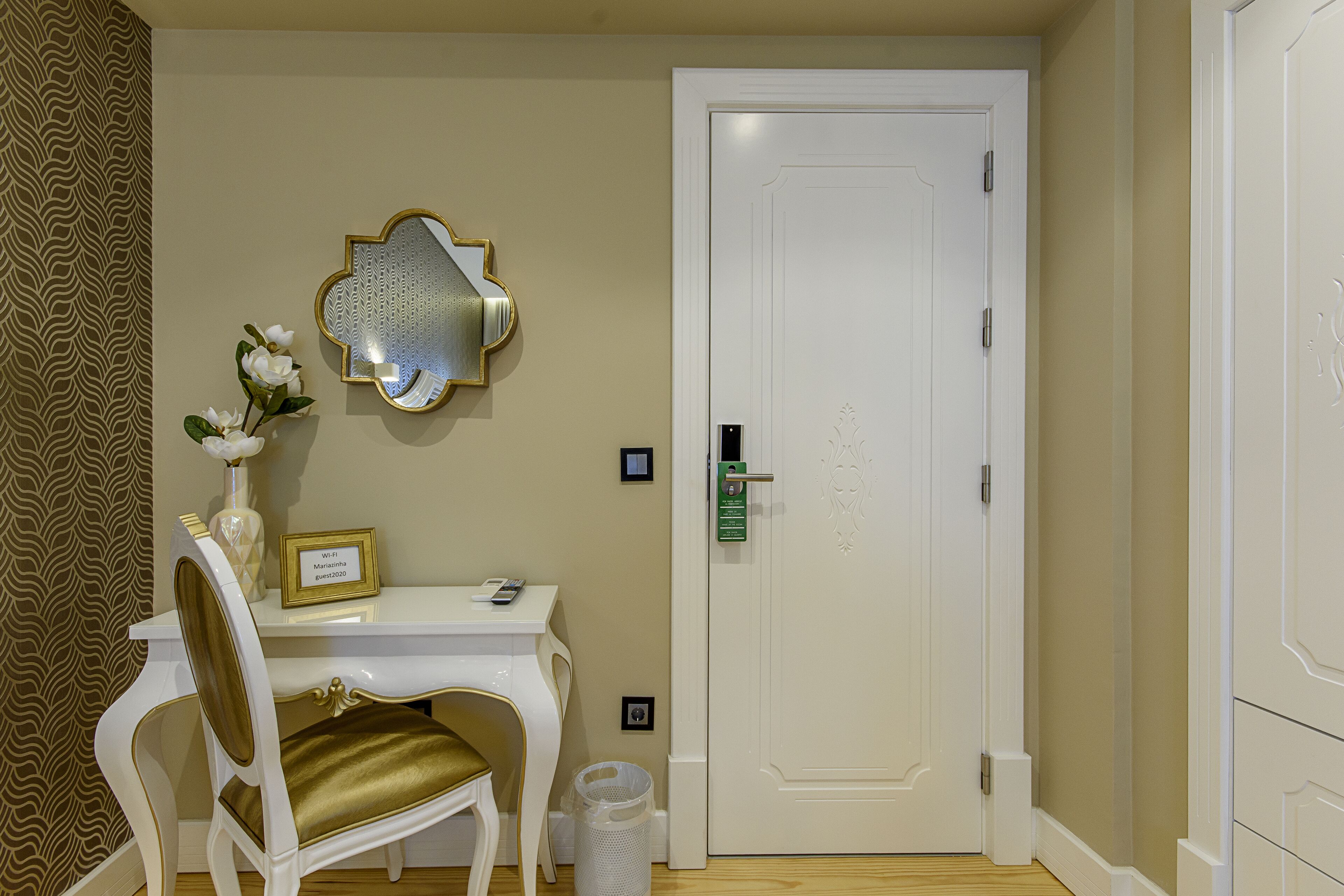 A hotel room with a white desk, a chair with a gold cushion, a mirror, and a door with a keycard lock.