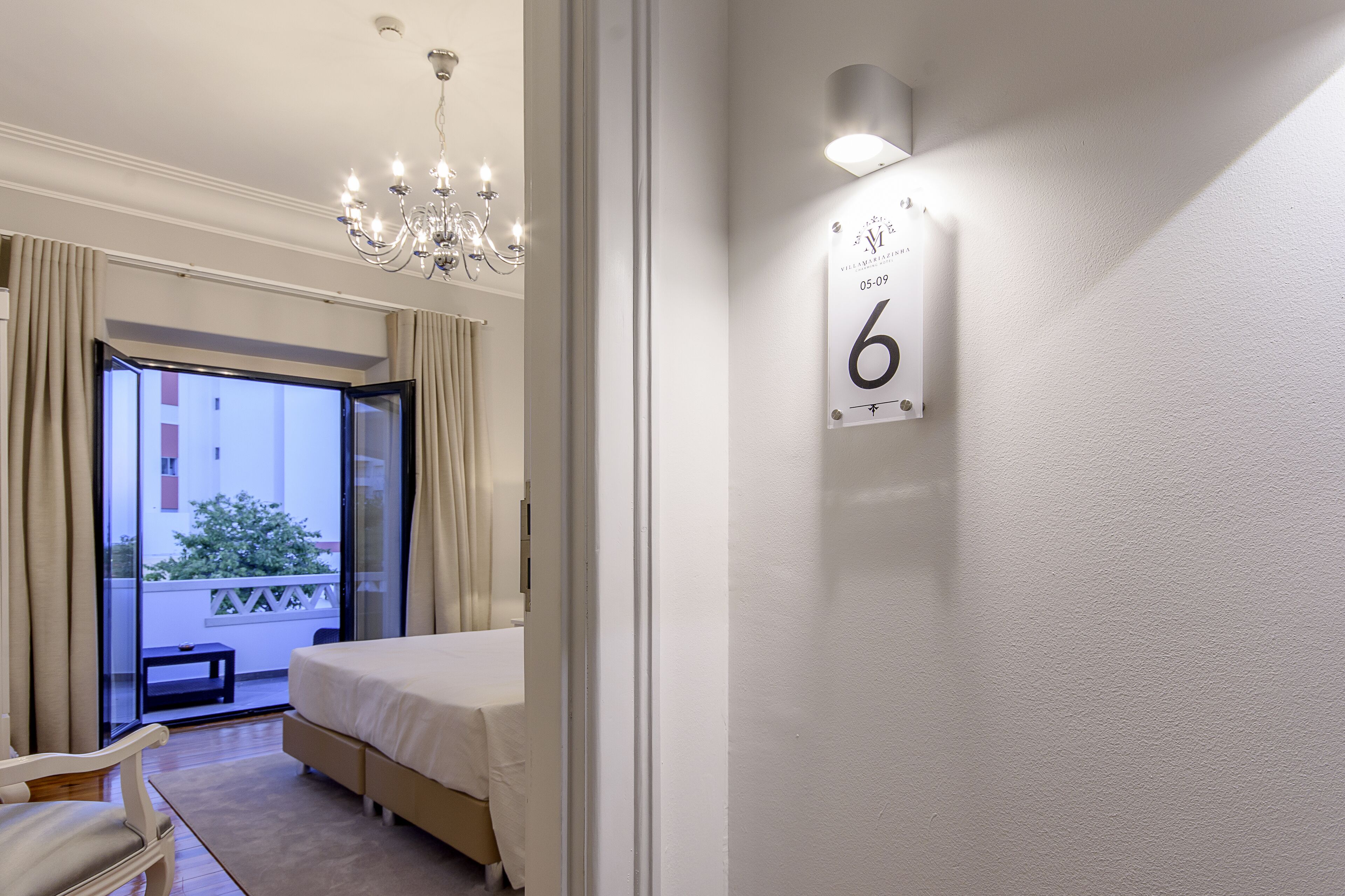 Hotellihuone, jossa on sänky, kattokruunu, parveke, jossa on verhot, ja seinälle kiinnitetty kyltti, jossa on numero 6.