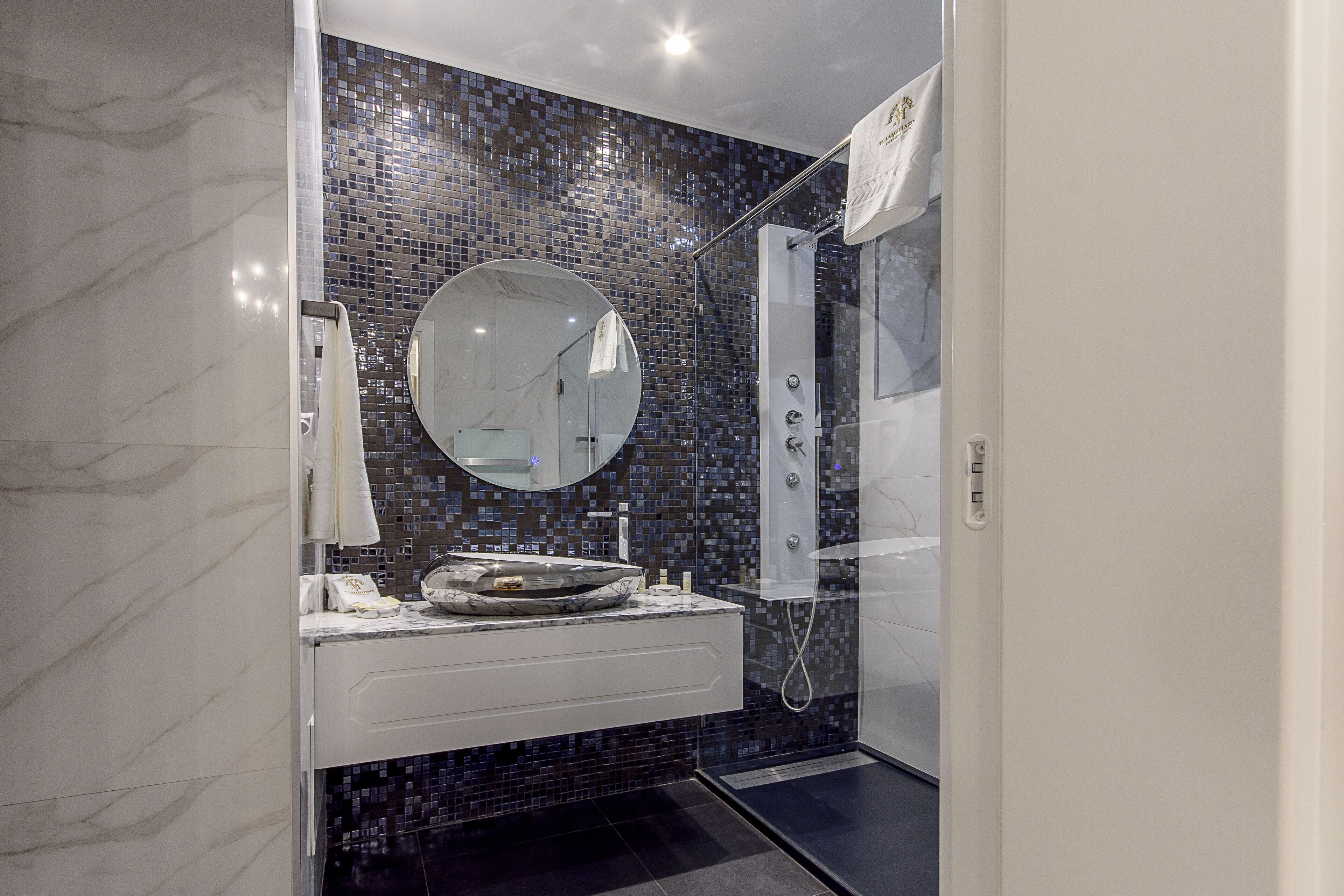 Un baño moderno con tocador de mármol, espejo redondo, cabina de ducha de vidrio y salpicadero de mosaicos.