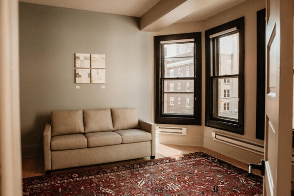 A room with a beige sofa, a patterned rug, and a large window with a view of buildings. - Dubbel Dutch (Milwaukee)