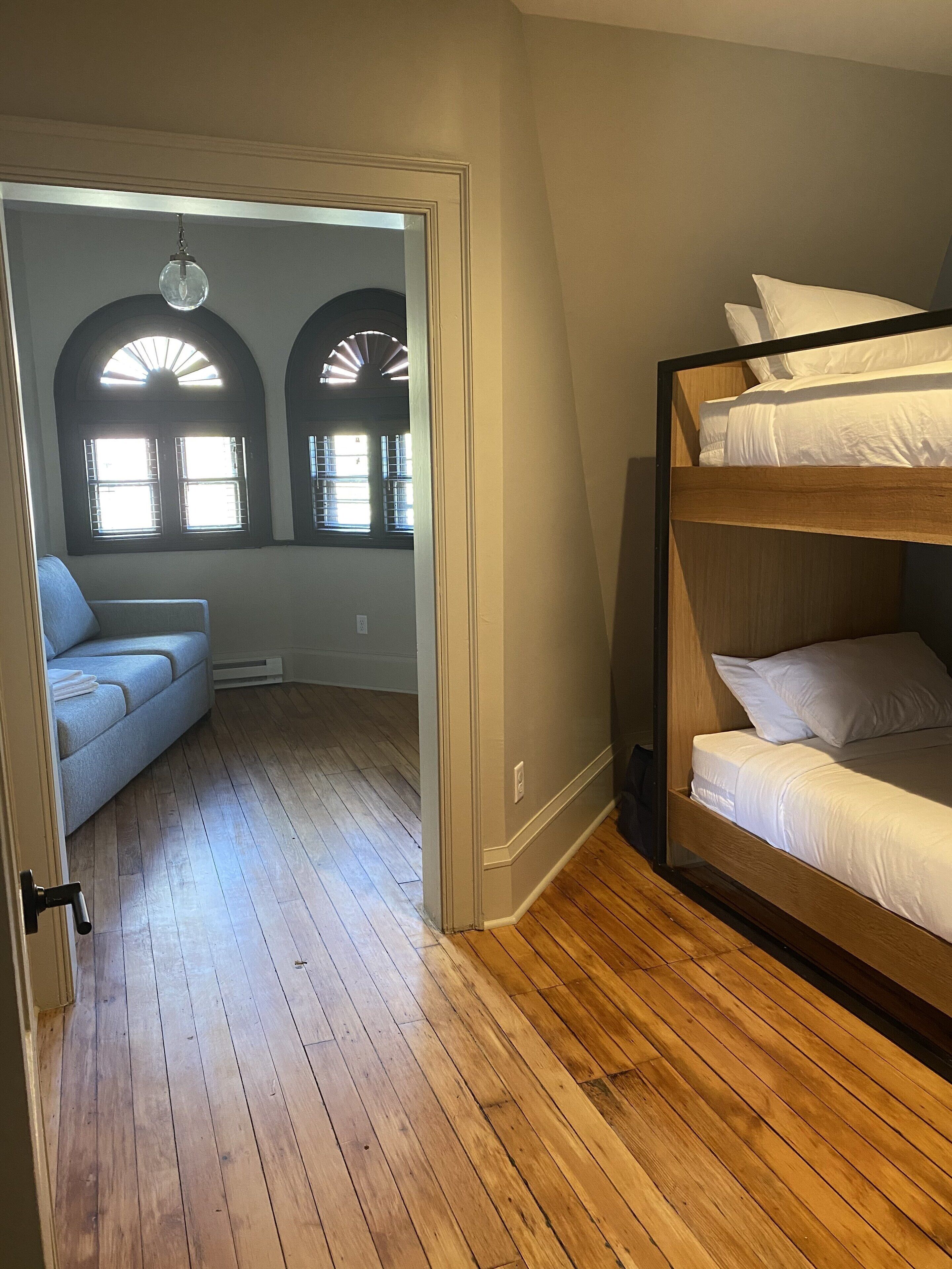 Een kamer met een stapelbed, een slaapbank en twee boogvensters met zonwering.