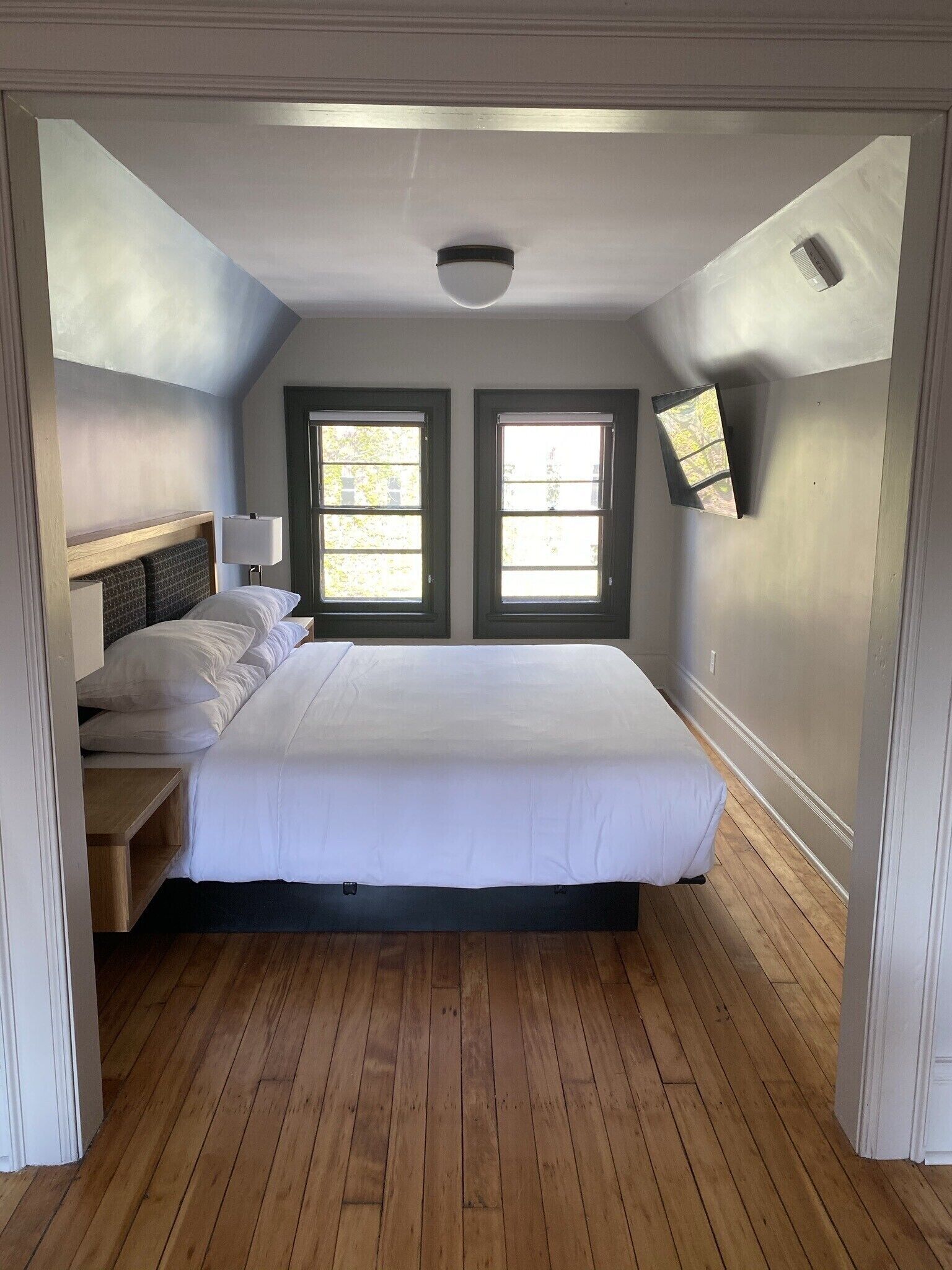 A bedroom with a large bed, two windows, a ceiling light, and a wall-mounted TV.