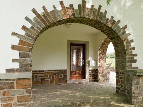 A stone archway leading to a door with a wooden frame, a stone-paved entrance, and a white wall with a small window.