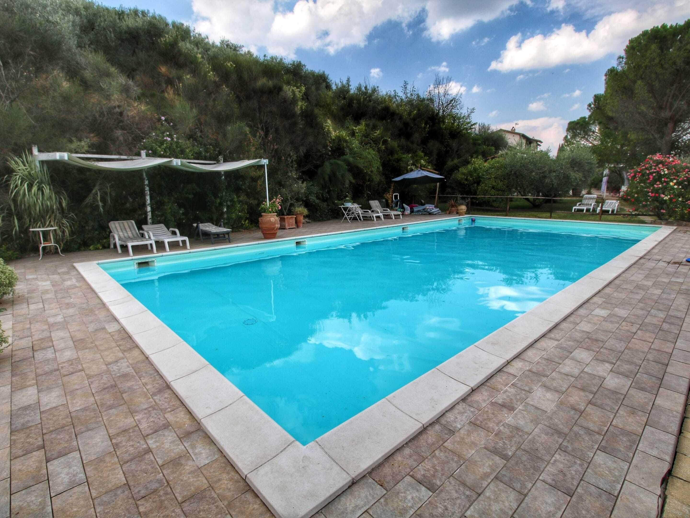 A rectangular swimming pool with a tiled edge, surrounded by a patio with stone tiles. There are lounge chairs and a covered seating area with a table and chairs.