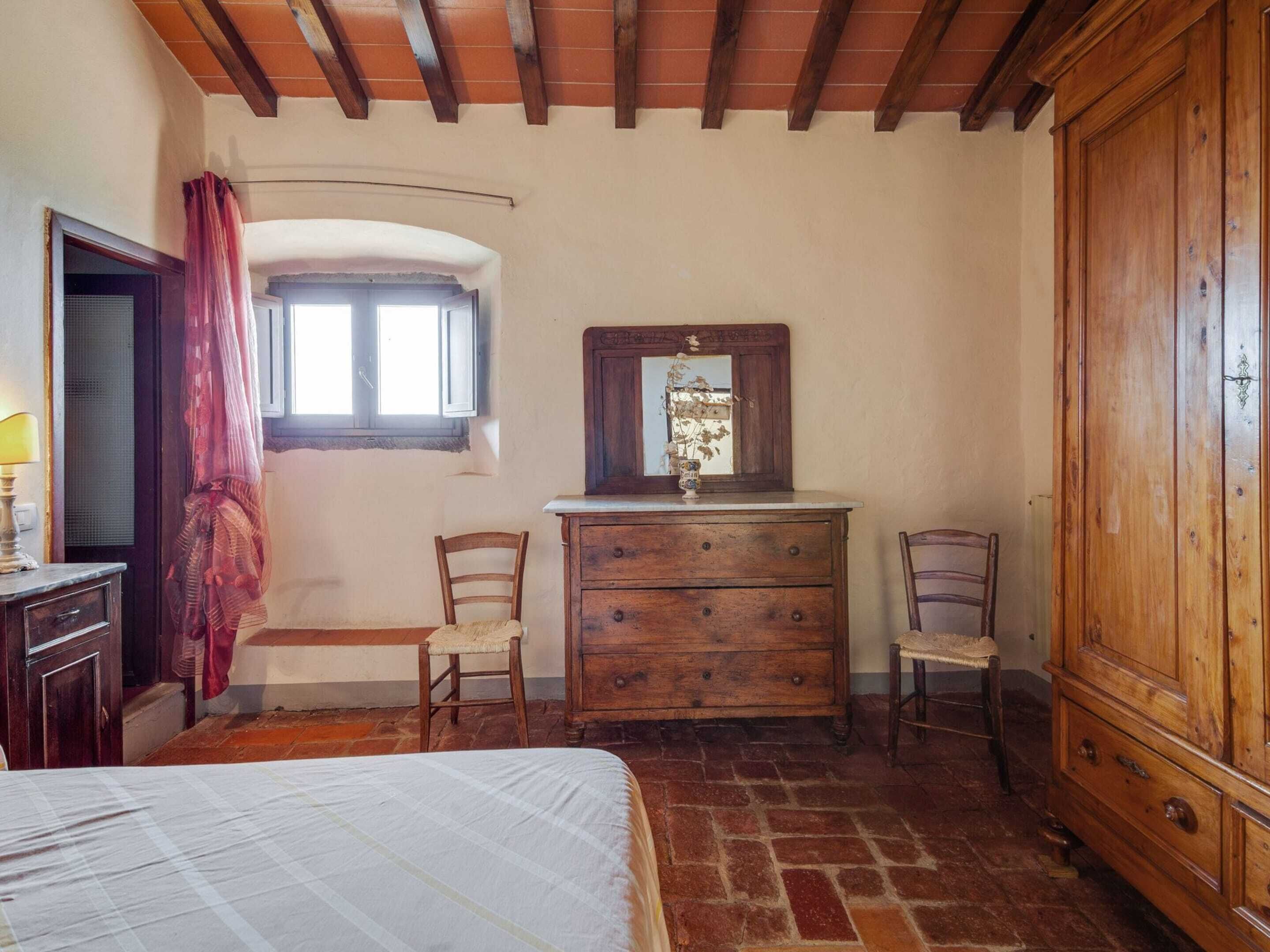 A bedroom with a bed, wooden dresser, two chairs, a window with curtains, and a mirror.