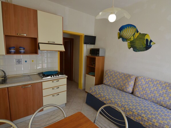 A compact kitchen with wooden cabinets, a gas stove, and a microwave. There's a sofa with a checkered pattern and a wall-mounted TV. - Belvilla by OYO Lido Estensi Bilo Doria (Comacchio)