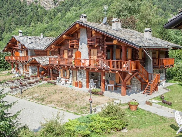 Une cabane en rondins dotée d’un balcon, avec des fondations en pierre et un paysage montagneux en arrière-plan.