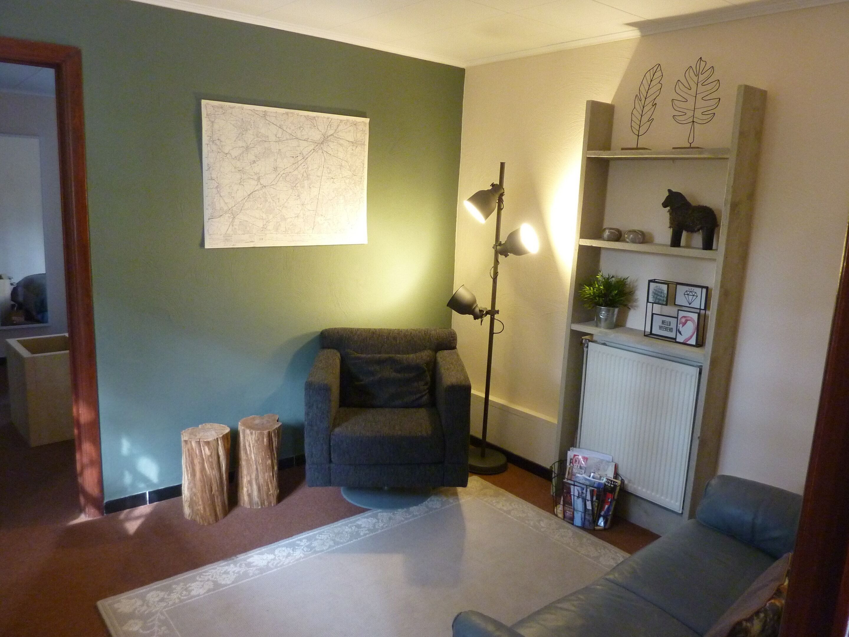 Una sala de estar con un sofá gris, una lámpara de pie, un mapa en la pared, una estantería con adornos y un radiador.