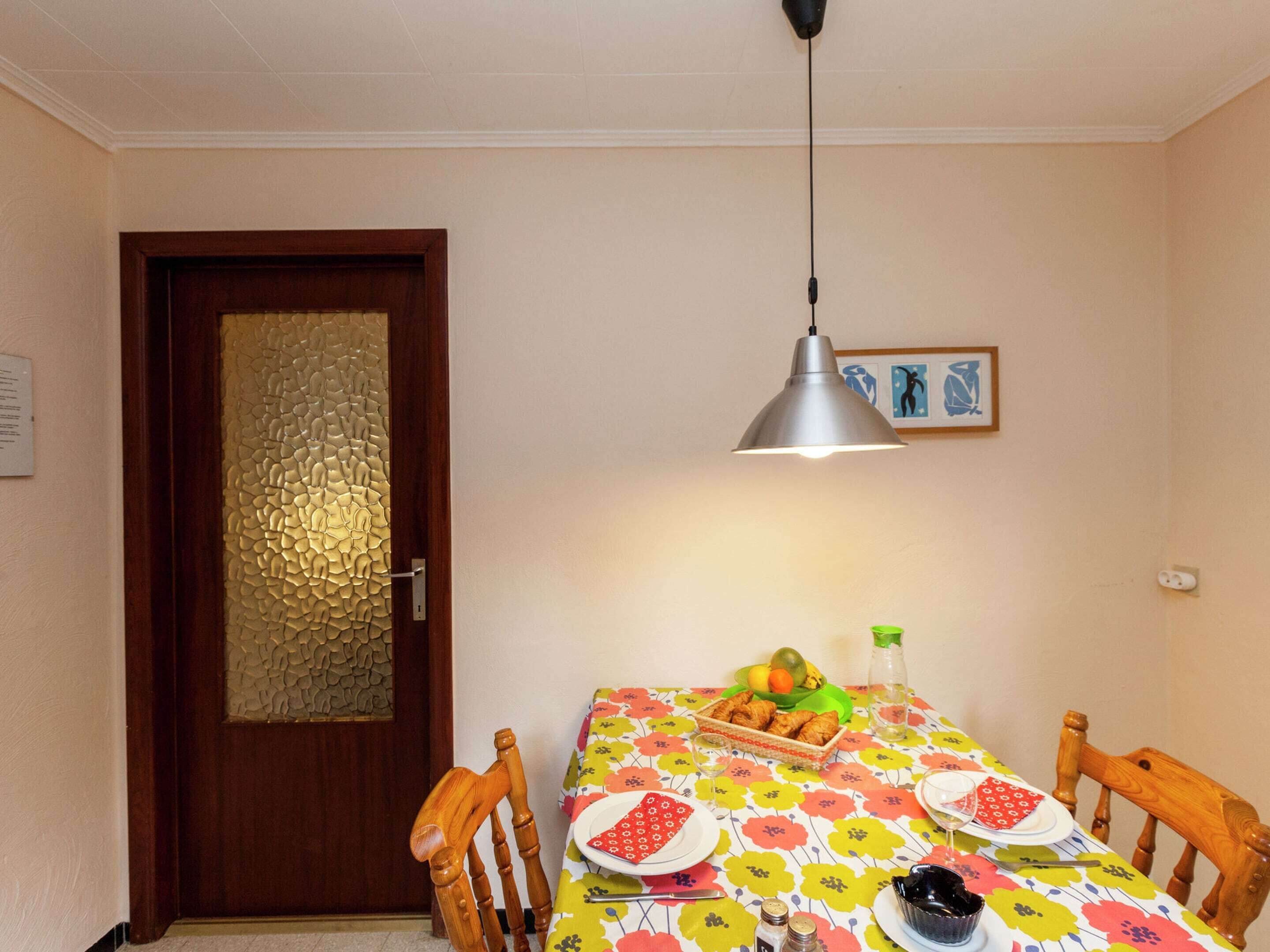 Un comedor con una mesa de madera, sillas, mantel estampado, cesta de frutas y una lámpara colgante.