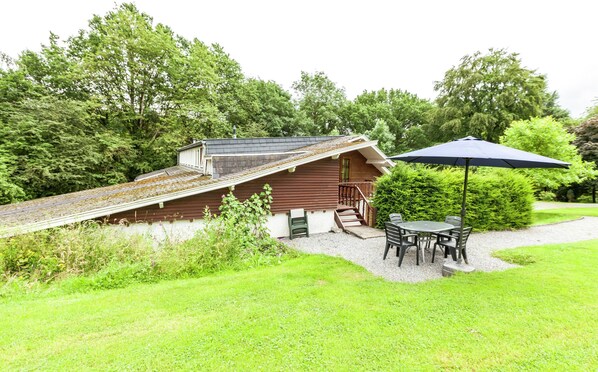 Een houten chalet met een schuin dak, een trap naar een veranda en een tafel met stoelen onder een blauwe parasol.
