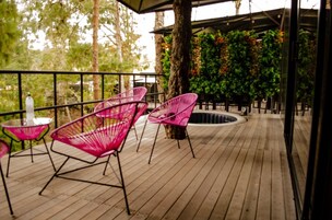 Una terraza de madera con sillas rosadas y una mesa de vidrio, rodeada de follaje y una pared decorativa.