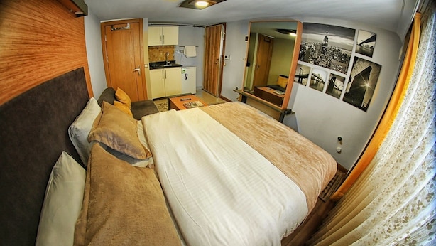 Une chambre compacte comprenant un lit, une petite kitchenette, un miroir et une photographie représentant un paysage urbain accrochée au mur.