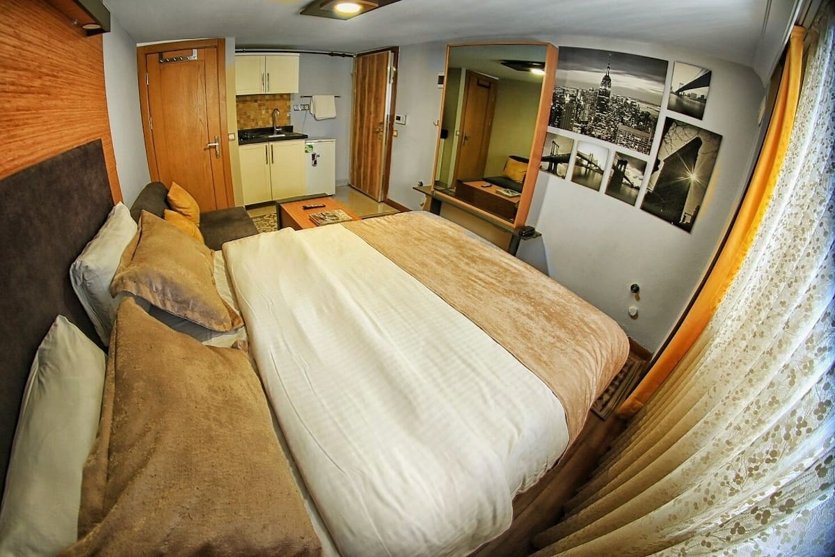 A compact bedroom with a bed, a small kitchenette, a mirror, and a cityscape photo on the wall.
