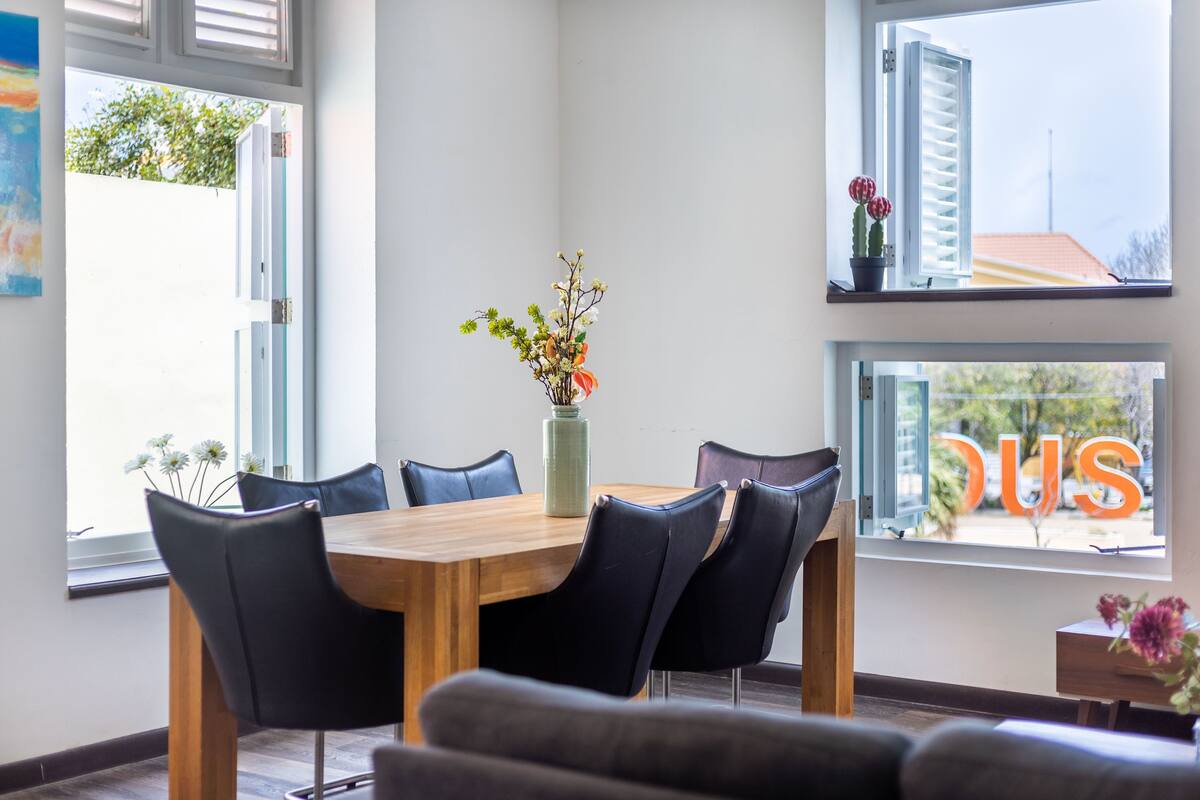 A dining area with a wooden table and black chairs, a vase with flowers, and a window with a view of a building and trees.