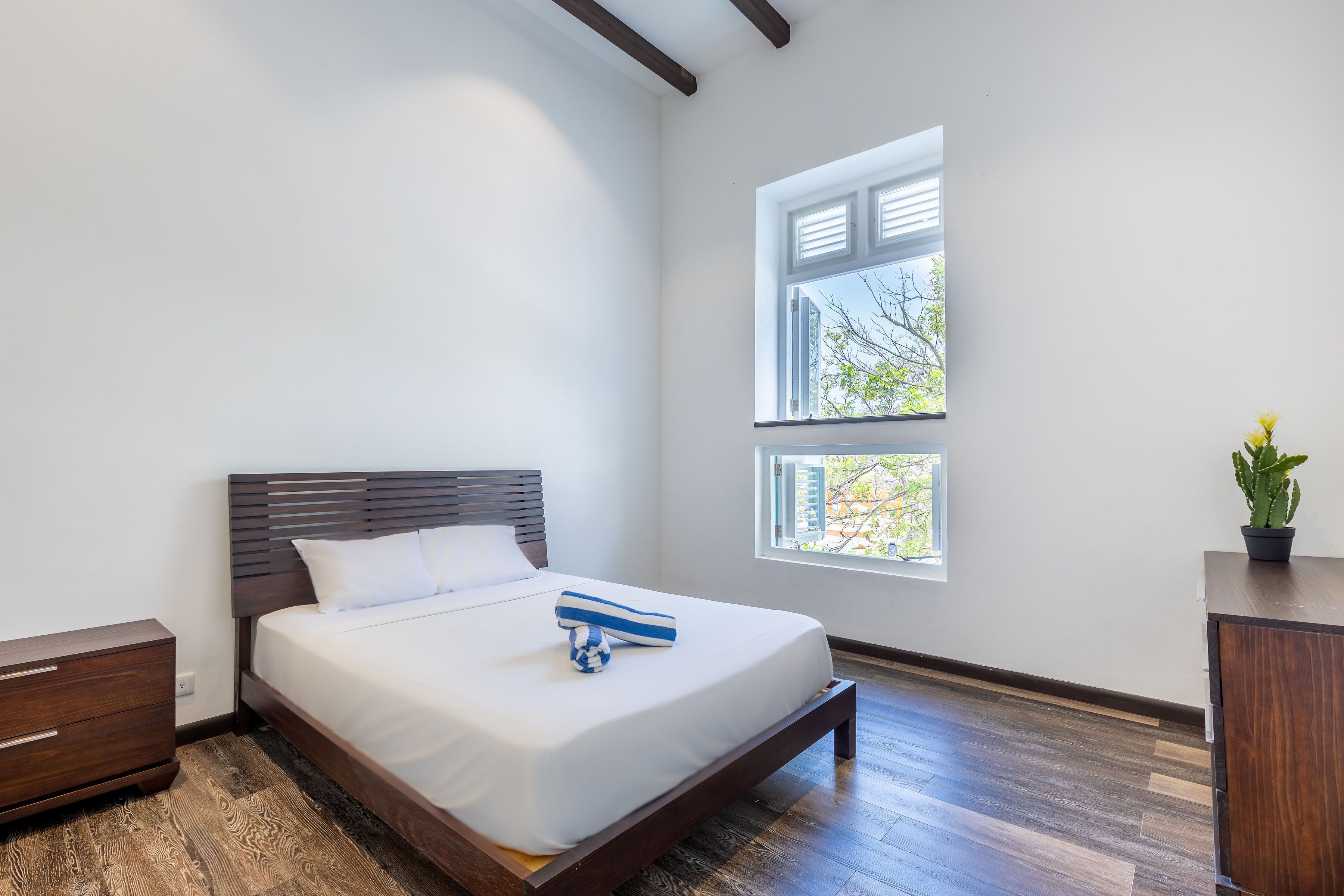 A bedroom with a wooden bed, white bedding, a nightstand, a dresser, and two windows with blinds.