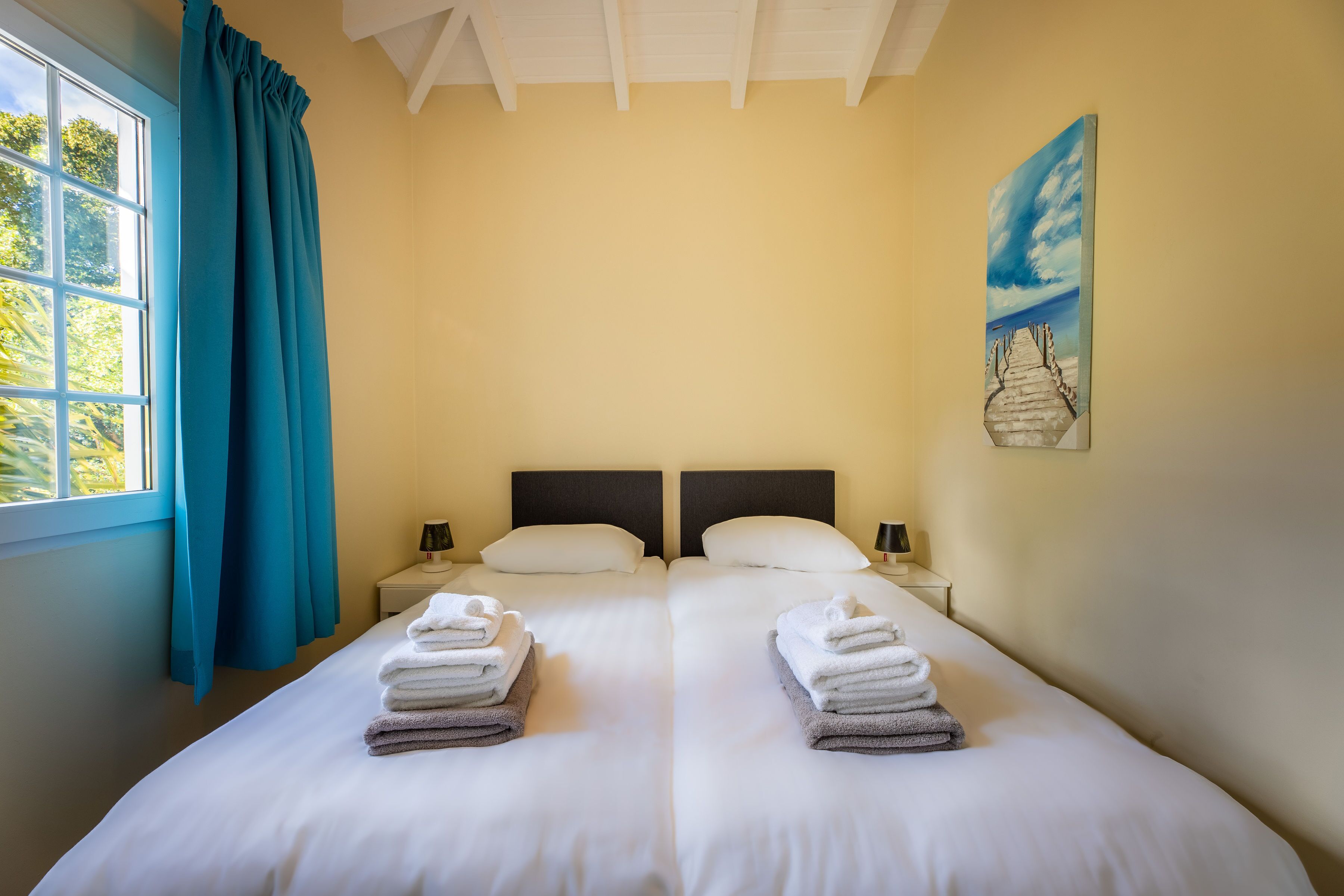 Un lit double avec du linge de lit et des serviettes blanches, une fenêtre avec des rideaux bleus et un tableau représentant une scène de plage accroché au mur.