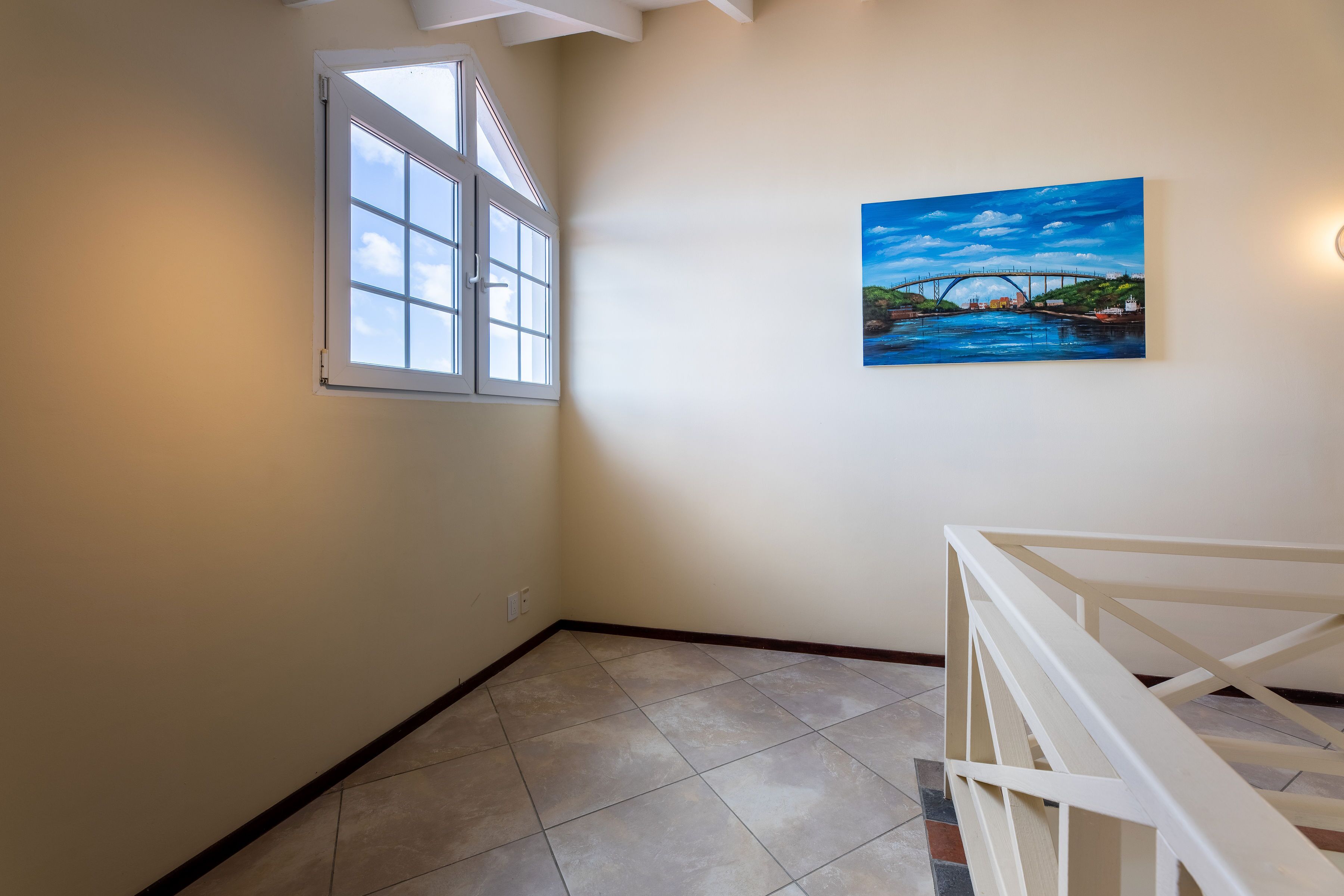 Una habitación con un ventanal grande, un cuadro de un puente y una barandilla blanca.