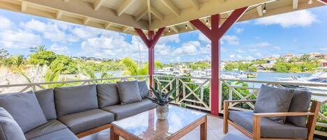 Un espacio de estar al aire libre cubierto con sofá, sillón y mesa de centro, con vista a un puerto deportivo con barcos y vegetación.