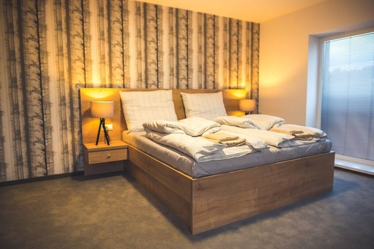 Quarto moderno com estrutura de cama de madeira, duas mesas de cabeceira com luminárias e uma janela grande com persianas.