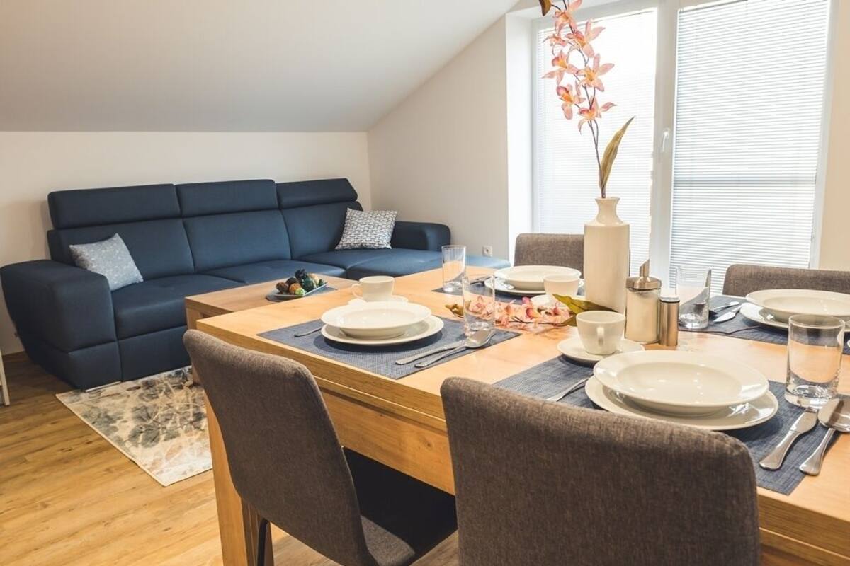 Uma sala de estar moderna com um sofá azul escuro, uma mesa de jantar de madeira posta com pratos, copos e talheres, e um vaso com flores.