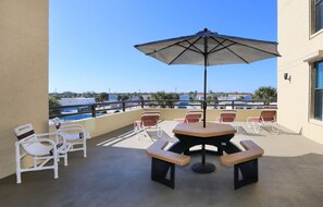 Relax on the balcony with a view of the pool and surrounding buildings, surrounded by comfortable lounge chairs and a shaded table.
