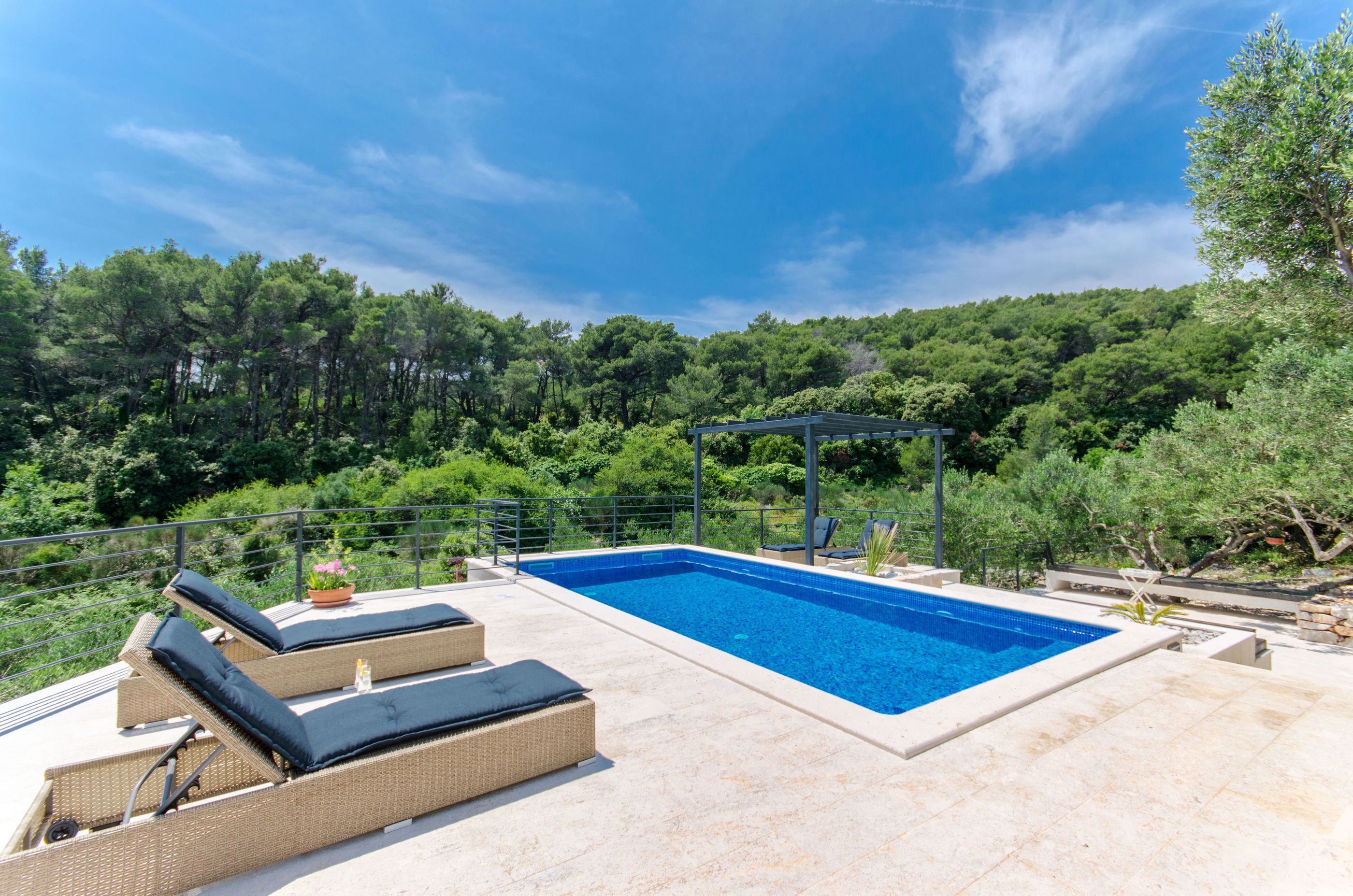 A rectangular swimming pool with lounge chairs and a pergola on a terrace surrounded by lush greenery.