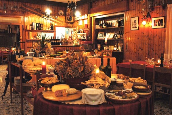A warmly lit dining area with wooden walls, a variety of food and drink items on tables, and a Christmas tree centerpiece.