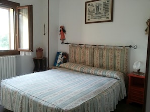 A bedroom with a bed, a window, a radiator, a small table with a lamp, and a framed picture on the wall.